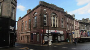 Colour photo of two-storey sandstone building, showing two sides. An estate estate agent's office occupies the ground floor; large windows indicating a more spacious and taller upper floor.