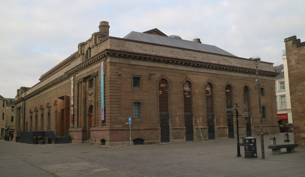 Colour photo of imposing 19th-century public building, two sides visible, with empty pedestrianised areas adjacent. Colourful banners hang from one side; tall narrow windows on the other.