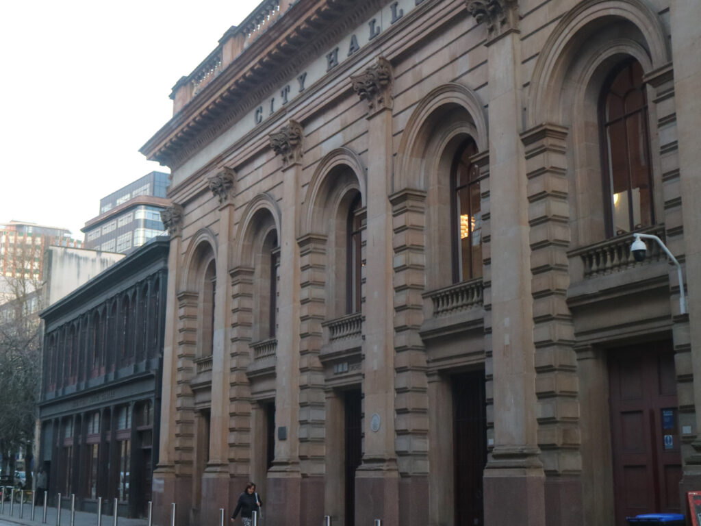 Imposing, renovated Victorian frontage of public building, identified in large letters under a decorated balcony as 'CITY HALLS'.