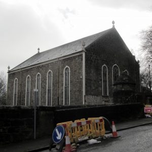 Photograph of church taken from across an adjacent road, two sides of the building visible beyond a retaining wall. Coned off roadworks in the foreground.